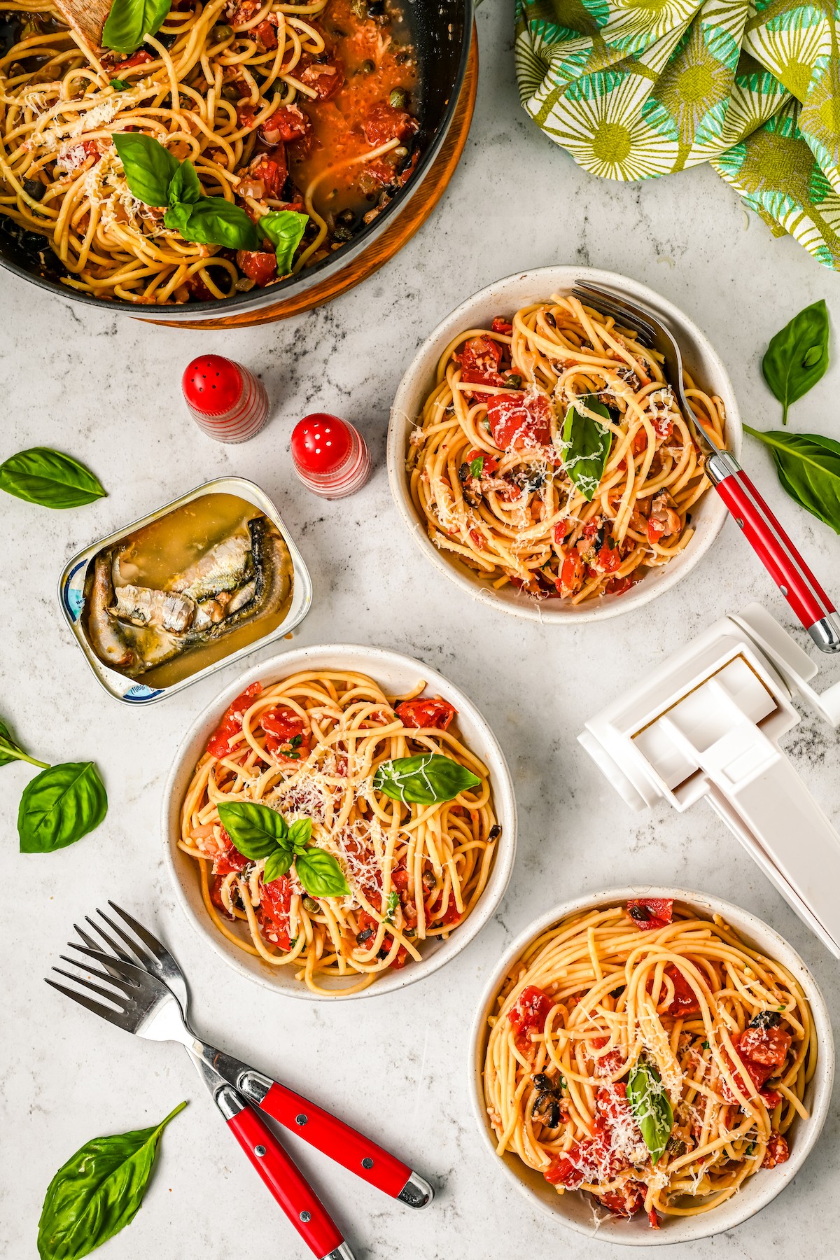 Bowls of pasta on a table with basil leaves, salt and pepper, and other ingredients.