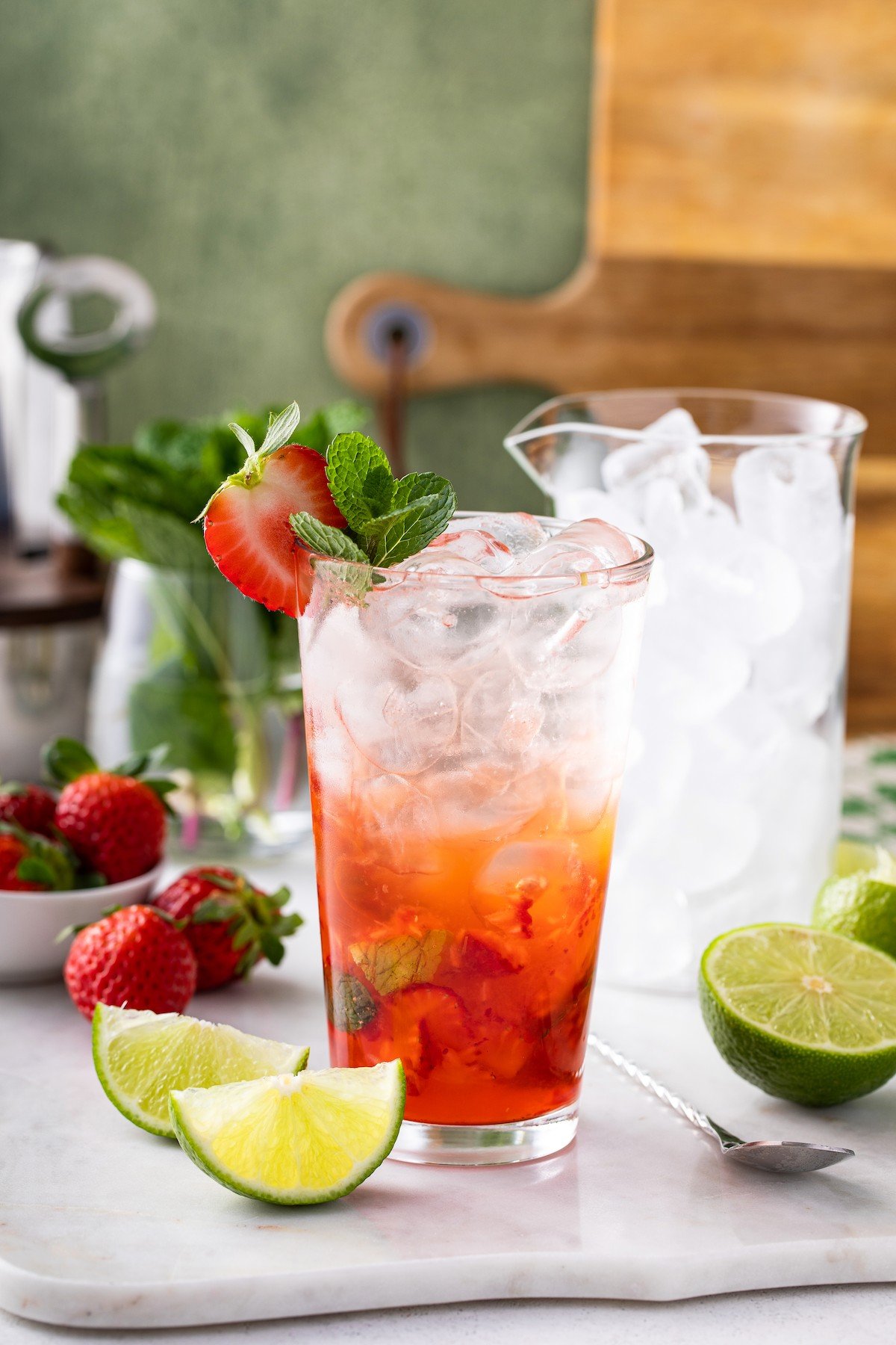 A strawberry mojito on a board next to a glass of ice, limes, and strawberries.