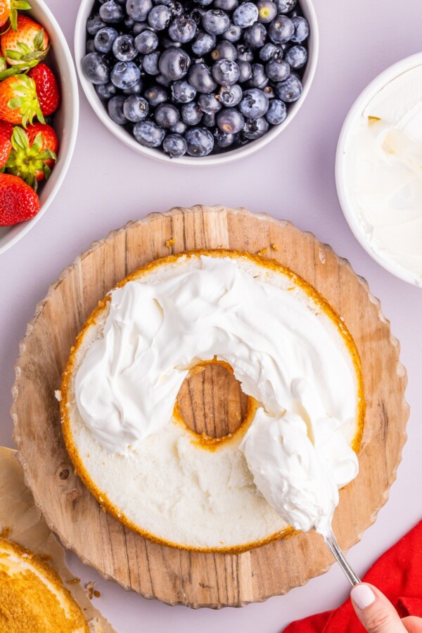 A layer of angel food cake topped with whipped cream. Bowls of strawberries, blueberries, and more whipped cream are on the work surface.