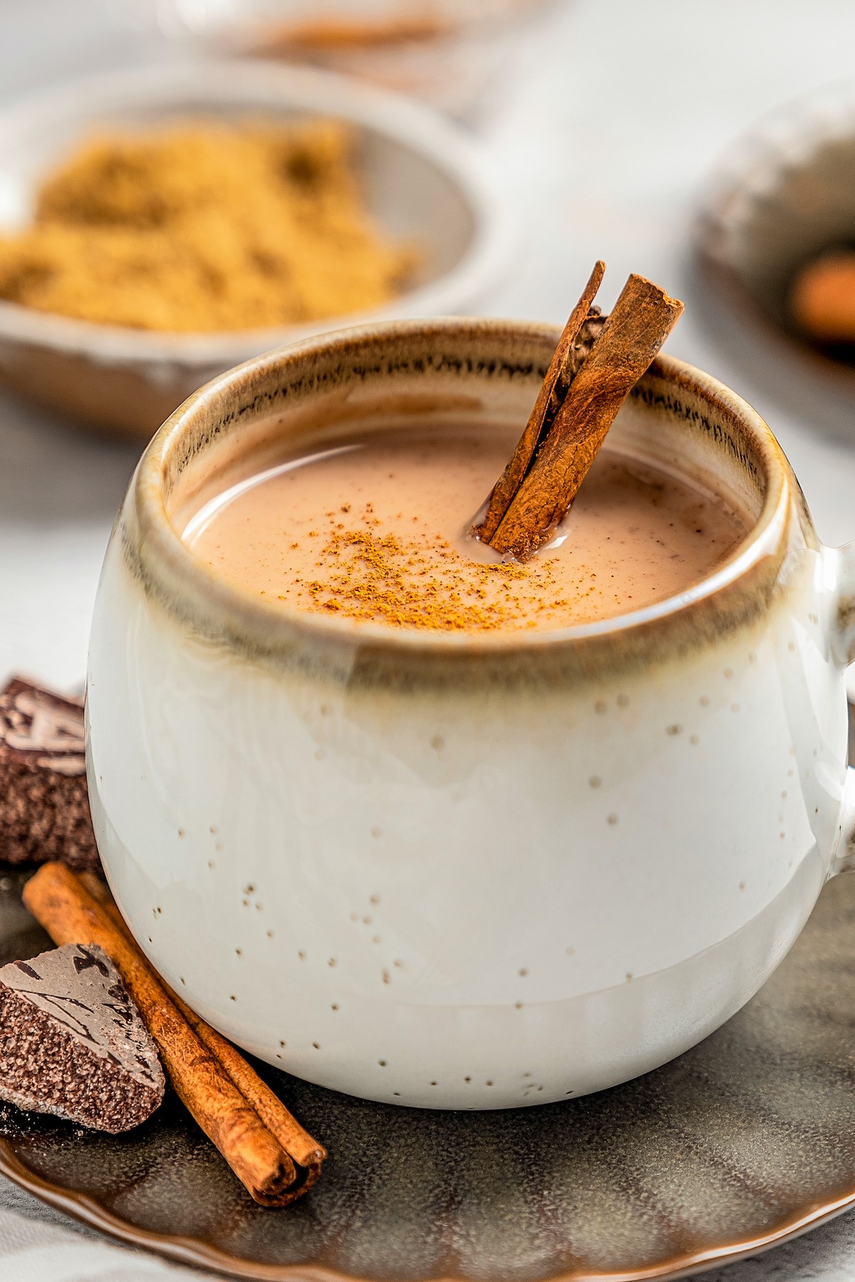 Creamy atole with chocolate in a mug with cinnamon sprinkled on top on a serving plate with cinnamon sticks.