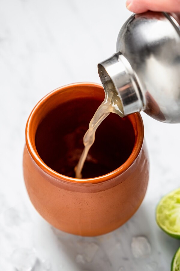 Cocktail shaker pouring shaken juice and tequila into a clay mug.