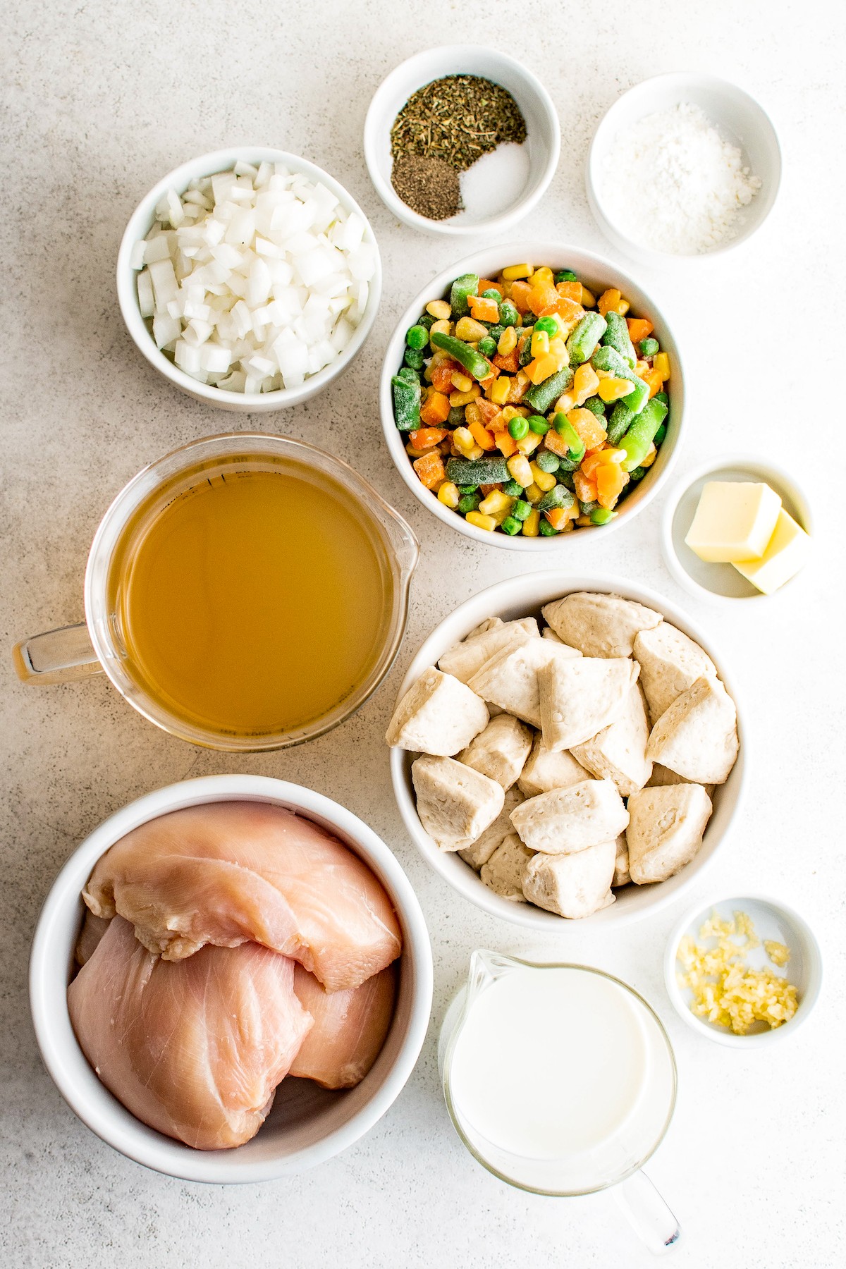 From top: Seasonings, cornstarch, frozen vegetables, butter, canned biscuits cut into pieces, garlic, evaporated milk, raw chicken breast, chicken broth, white onion.