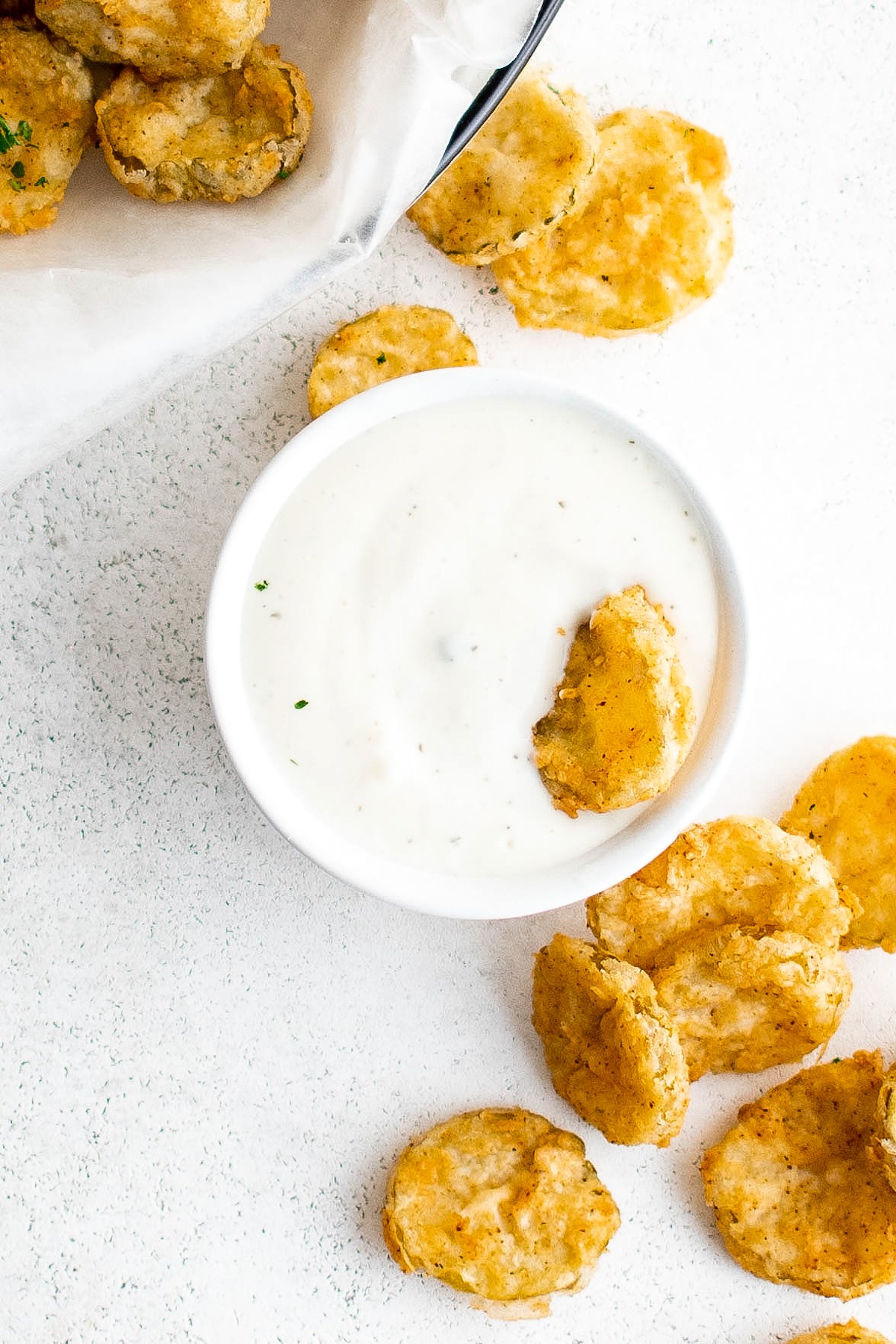 Fried pickles on a white table with a cup of ranch. One pickle is halfway dunked in the ranch.