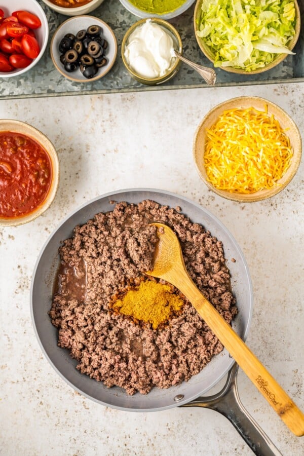 Ground beef in a skillet with taco seasoning. A dish of salsa and other taco ingredients are nearby.