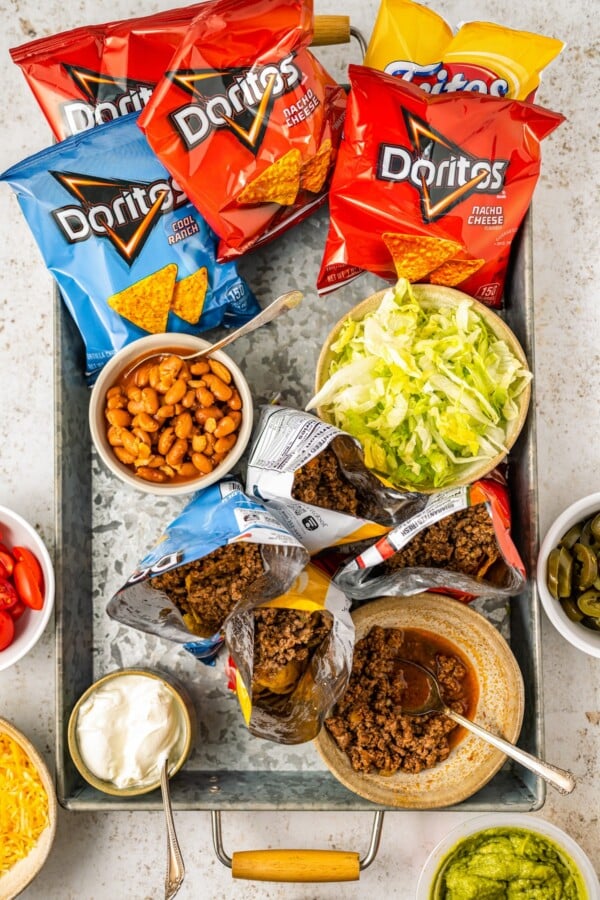 A tray of ingredients, including shredded lettuce, seasoned beef, beans, and chips. Some of the bags of chips are open and have beef added.