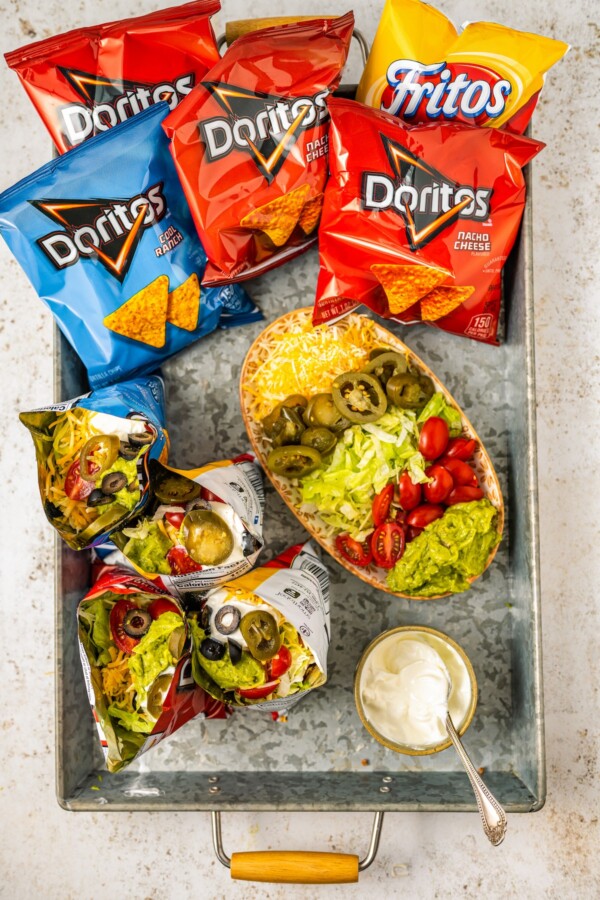 Four bags of chips that have been opened with toppings added, including sour cream, lettuce, and black olives. More toppings are arranged on the work surface.