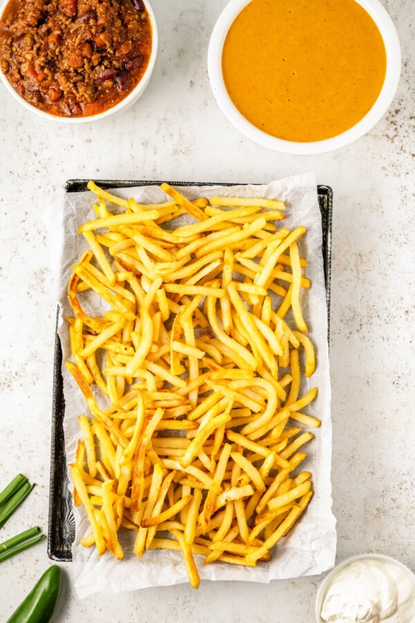A sheet pan of baked fries next to a bowl of seasoned cheese sauce and a bowl of chili.
