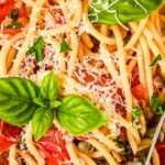 A bowl of Spaghetti Puttanesca with freshly shredded Parmesan on top and a fork tucked into the side of the bowl.