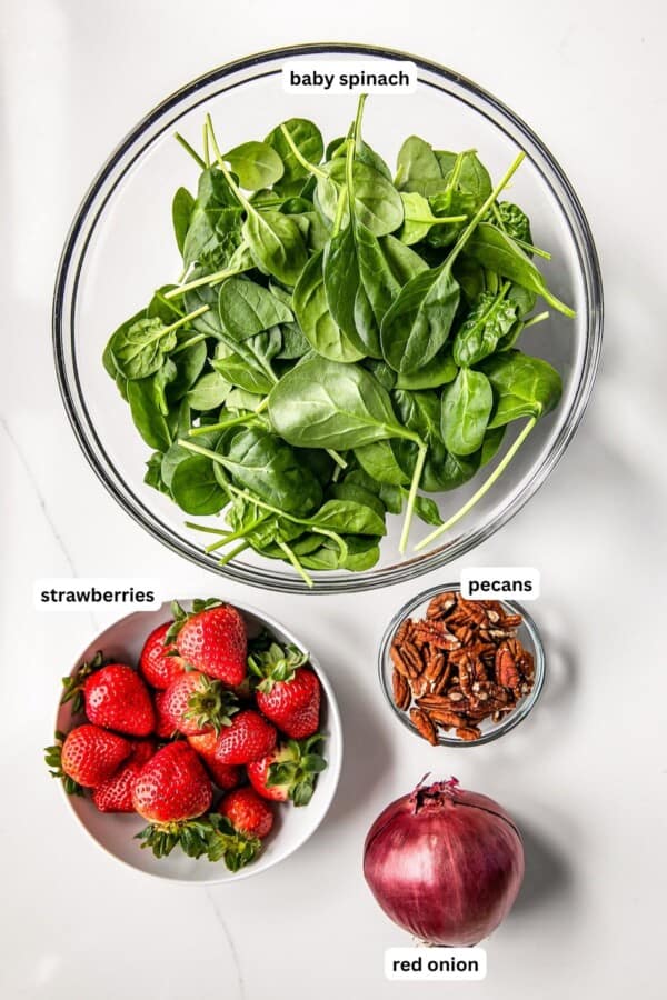 Ingredients for spinach salad recipe in order from top to bottom: baby spinach, strawberries, pecans and red onion.