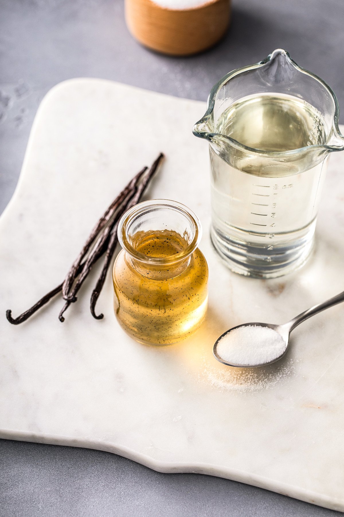 A jar of vanilla syrup next to a cup of water, a spoonful of sugar, and several whole vanilla beans.