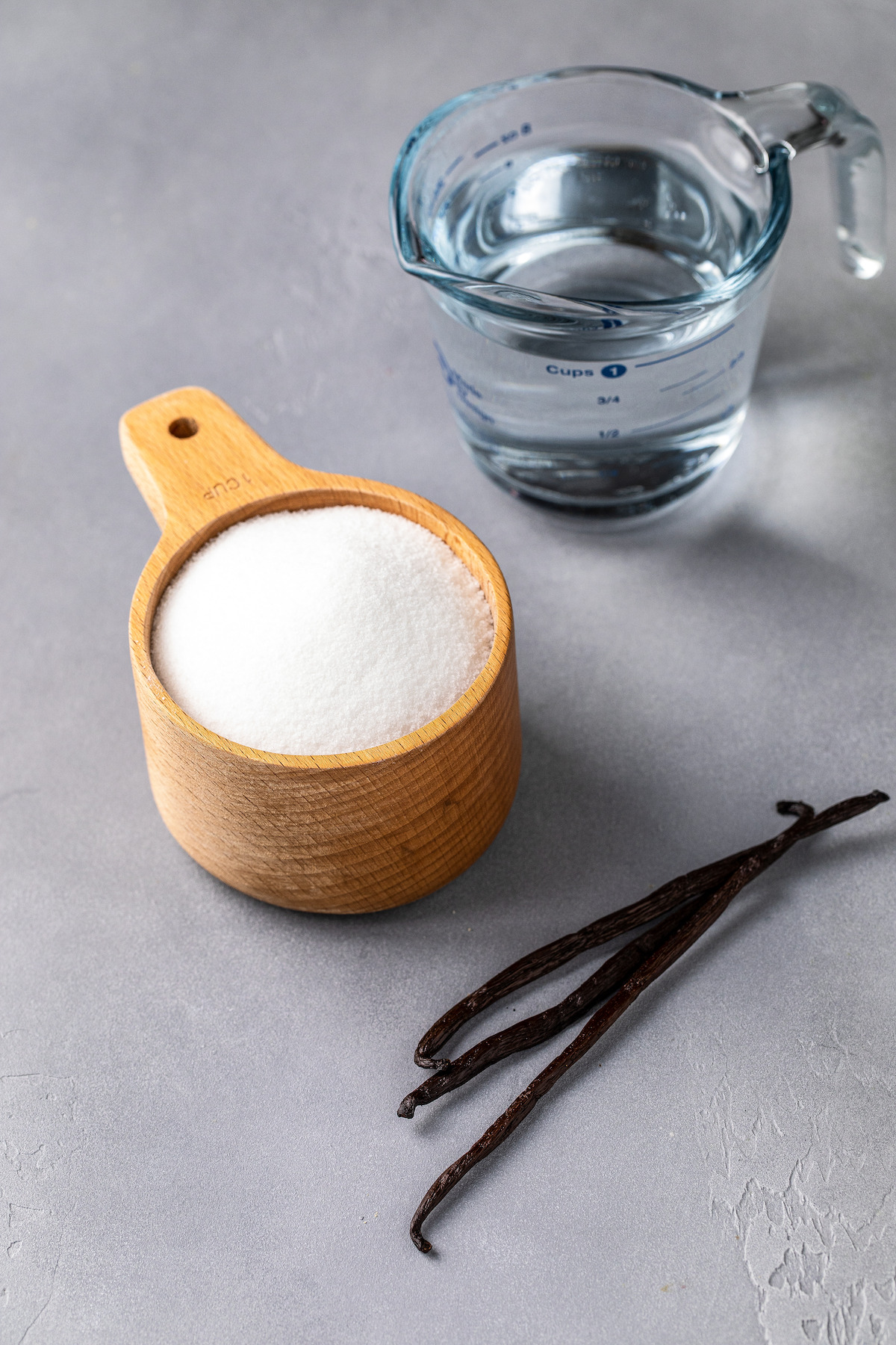 A measuring cup of water, a measuring cup of sugar, and three vanilla beans.