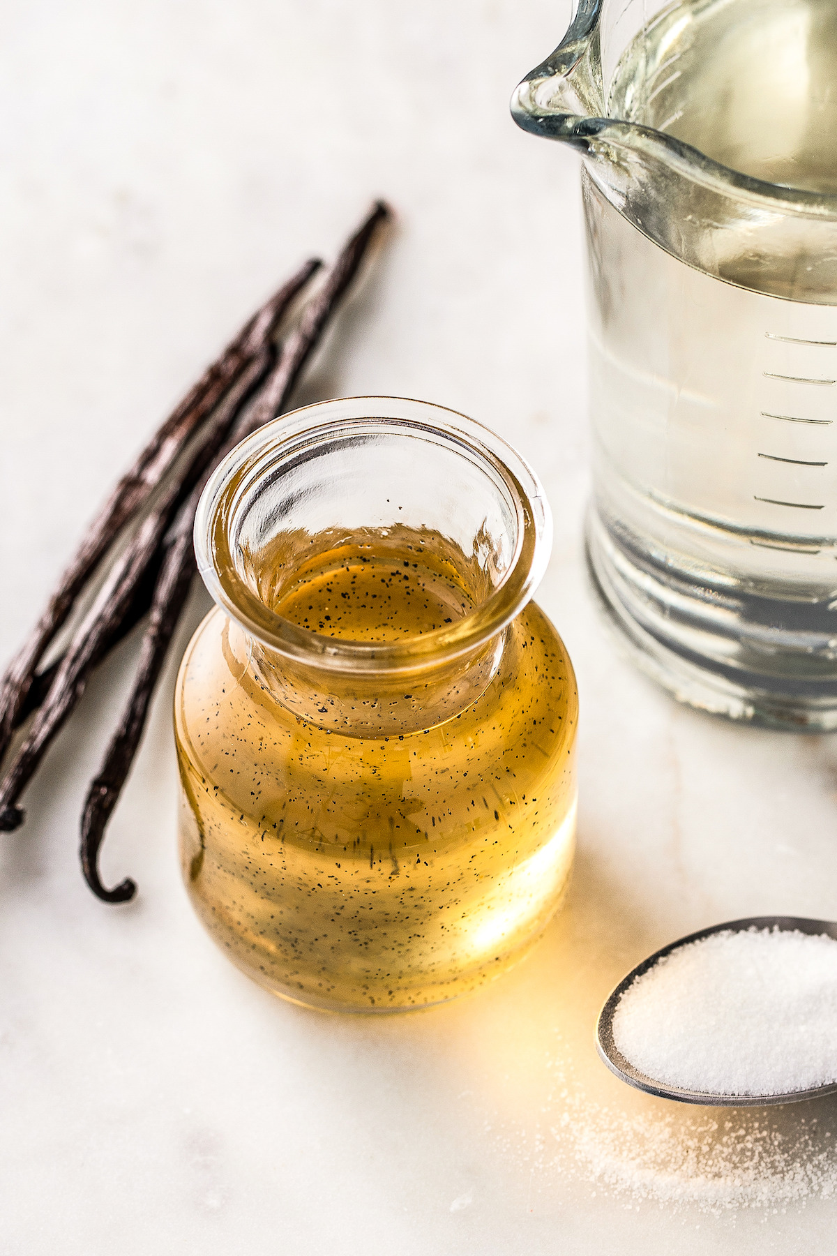 A measuring cup of water next to vanilla beans, sugar, and a jar of syrup.