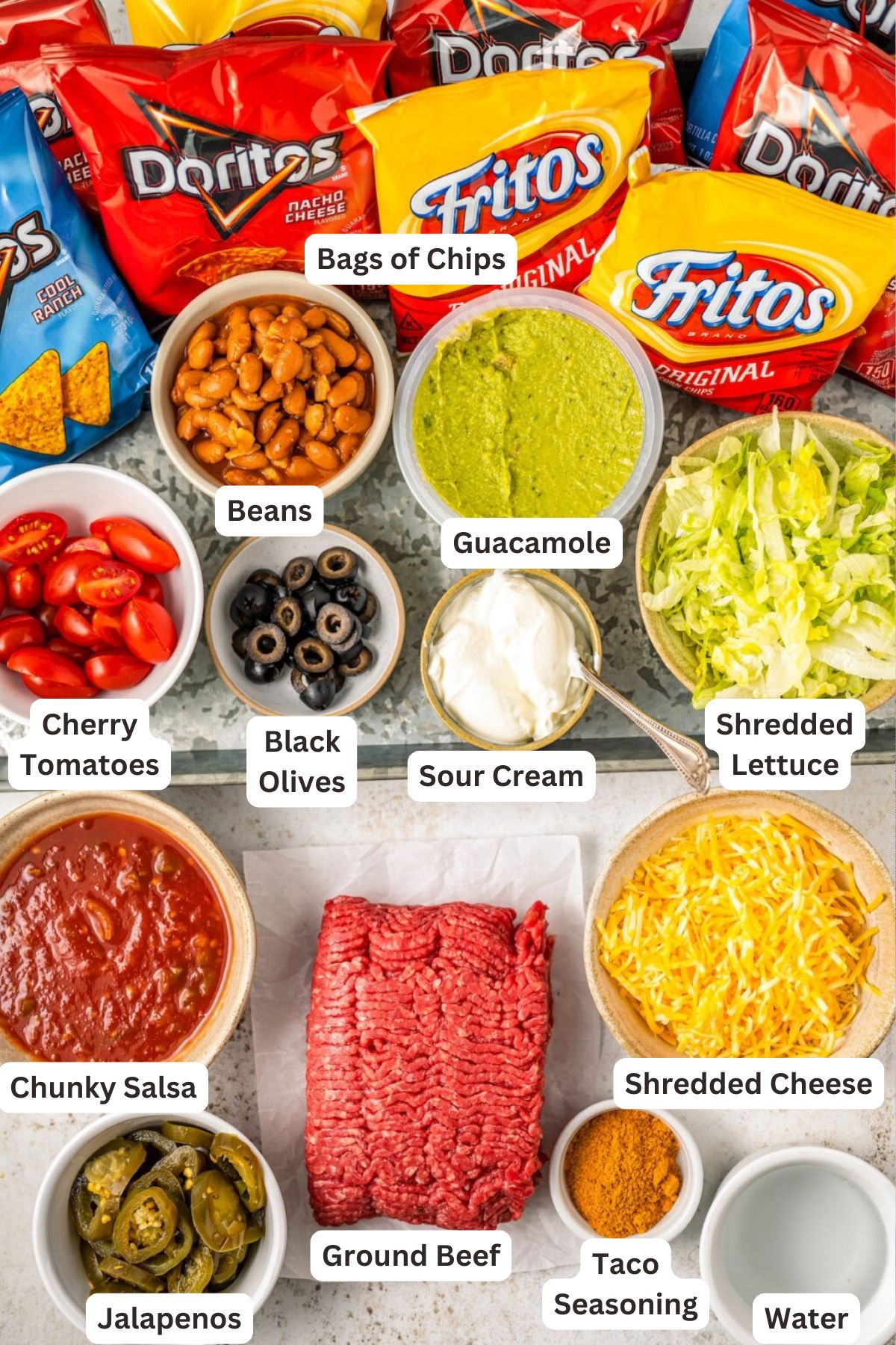 Ingredients for Walking Taco Recipe in order from top to bottom: bags of chips, beans, guacamole, cherry tomatoes, black olives, sour cream, shredded lettuce, chunky salsa, ground beef, shredded cheese, jalapenos, taco seasoning and water. 