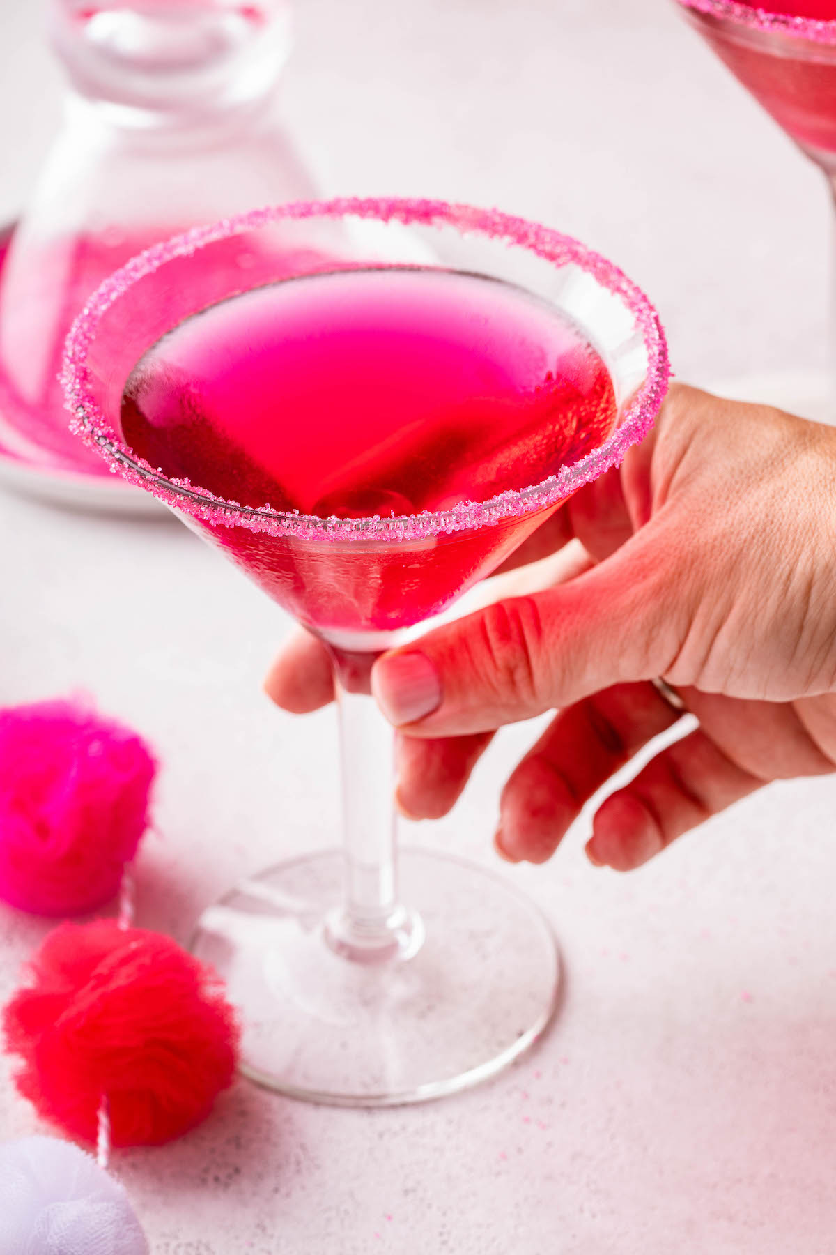 A hand picking up a pink martini with a pink sugar rim.