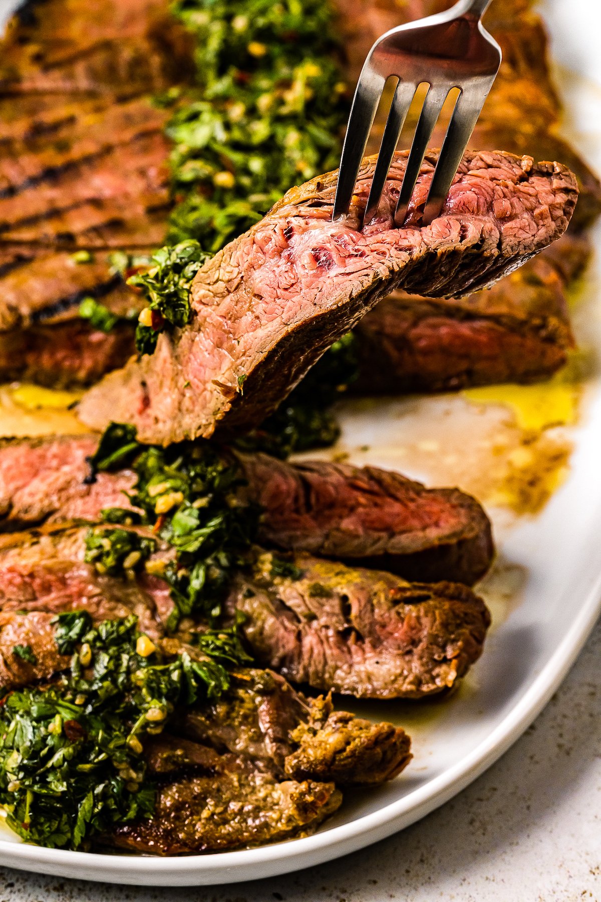 Sliced Churrasco Steak with chimichurri on top on a plate with a fork picking up a piece.