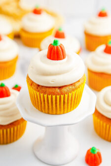 Pumpkin cupcakes lined up on a white surface. One is on a tiny cake stand.