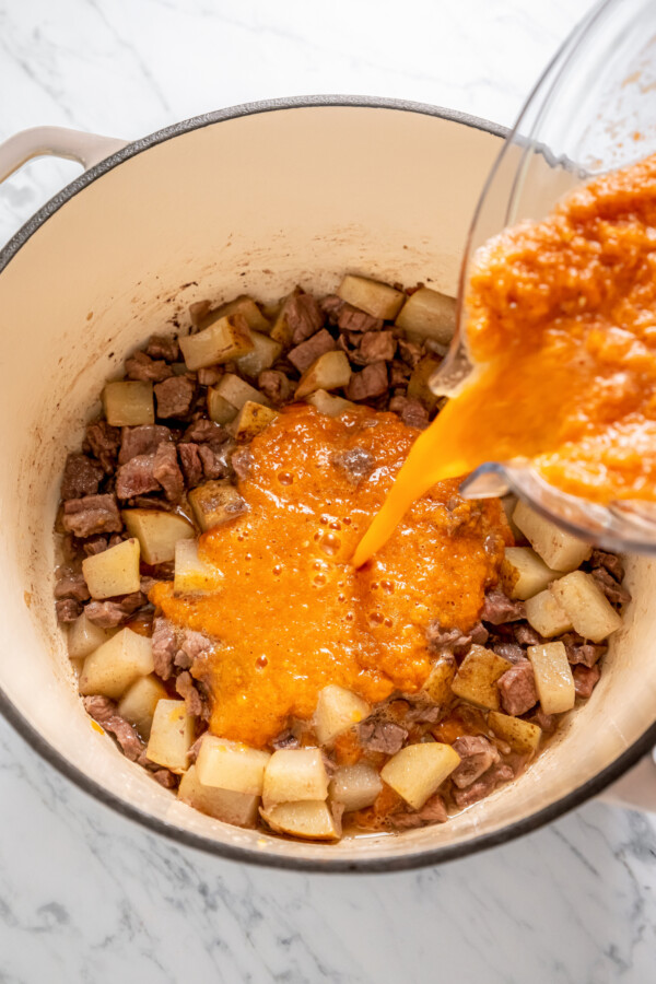 Pureed tomato sauce being poured into a pot with cubed potatoes and beef.