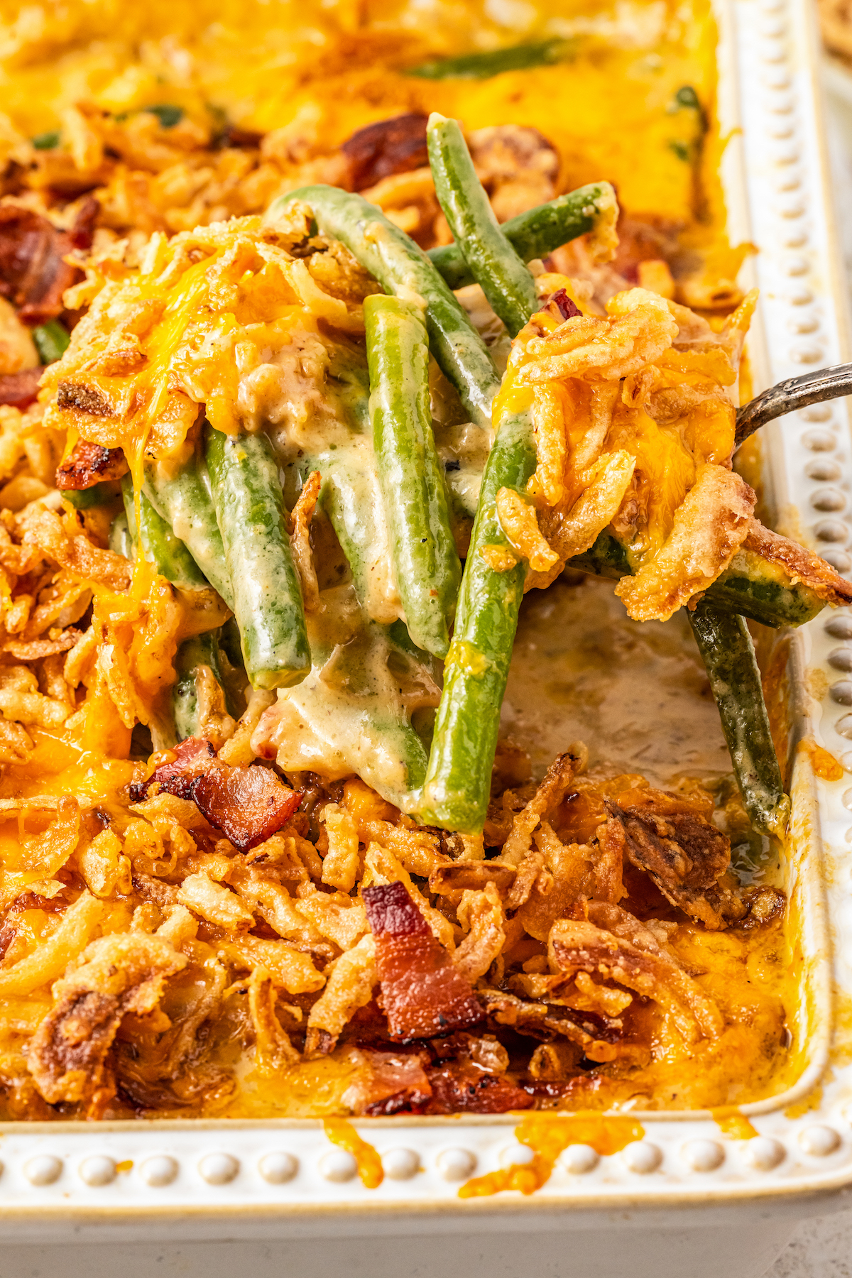 A serving spoon lifting a serving of green bean casserole with cheese out of a glass dish.