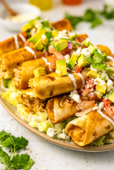 A platter of chicken taquitos on a bed of lettuce and topped with avocado, Mexican crema, tomatoes, onion, and crumbled cheese.