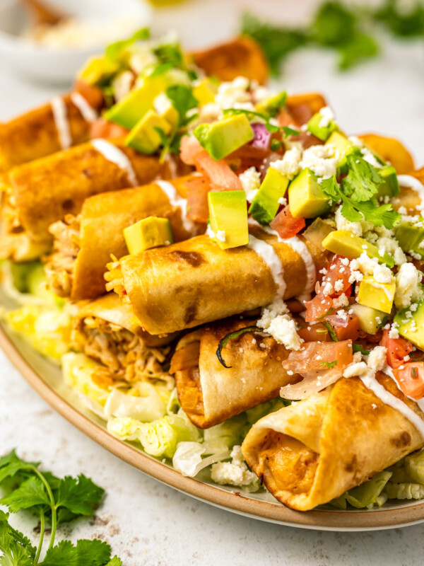 A platter of chicken taquitos on a bed of lettuce and topped with avocado, Mexican crema, tomatoes, onion, and crumbled cheese.