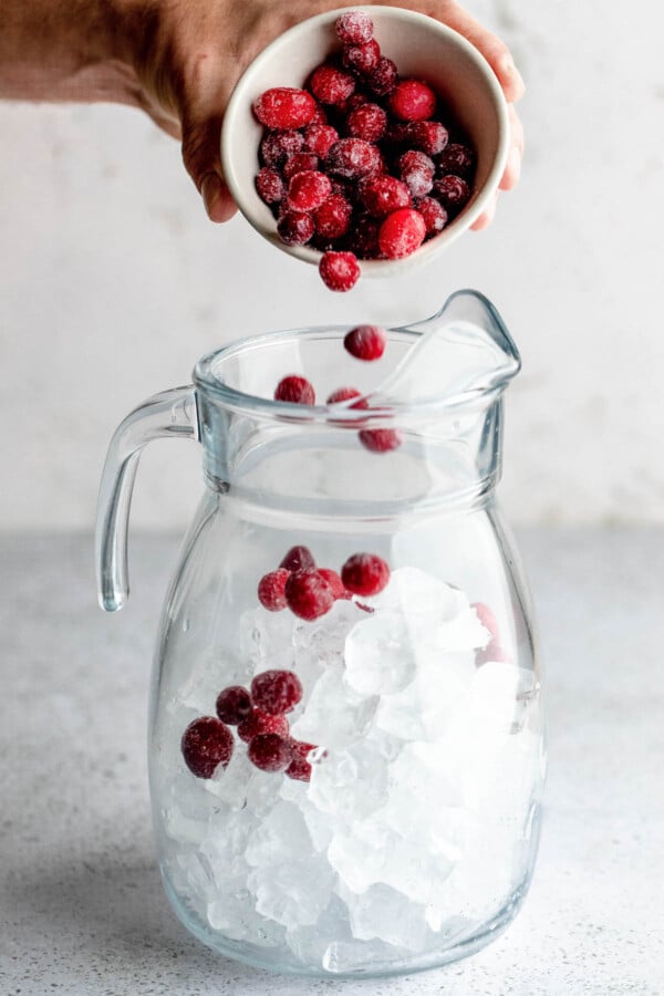 Sprinkling frozen cranberries into a pitcher with ice.
