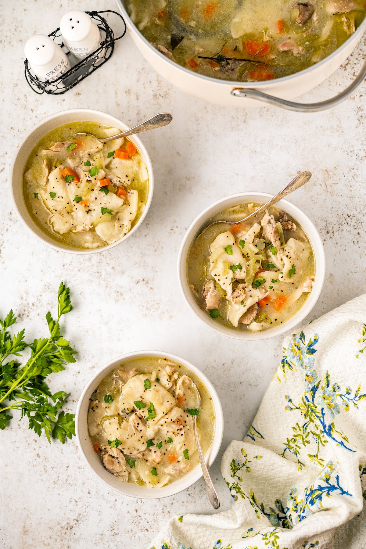 Three bowls of southern style chicken and dumplings on a table with fresh herbs and a cloth napkin.