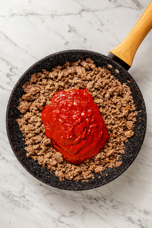 Chile sauce being added to a skillet with cooked ground beef.