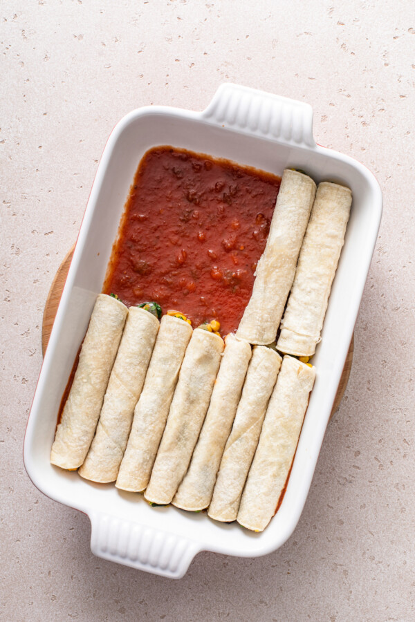 Enchiladas being filled and placed in a casserole dish filled with enchilada sauce.