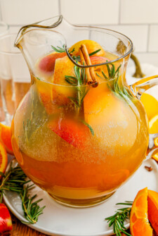 A pitcher of punch with apples, oranges, cinnamon, and rosemary.