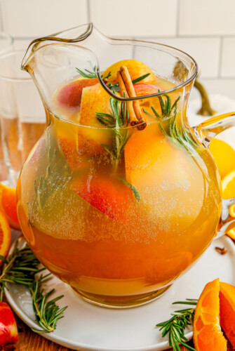 A pitcher of punch with apples, oranges, cinnamon, and rosemary.