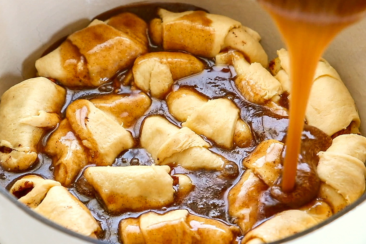 Apple cider sauce being poured over the top of apple dumplings with crescent rolls.
