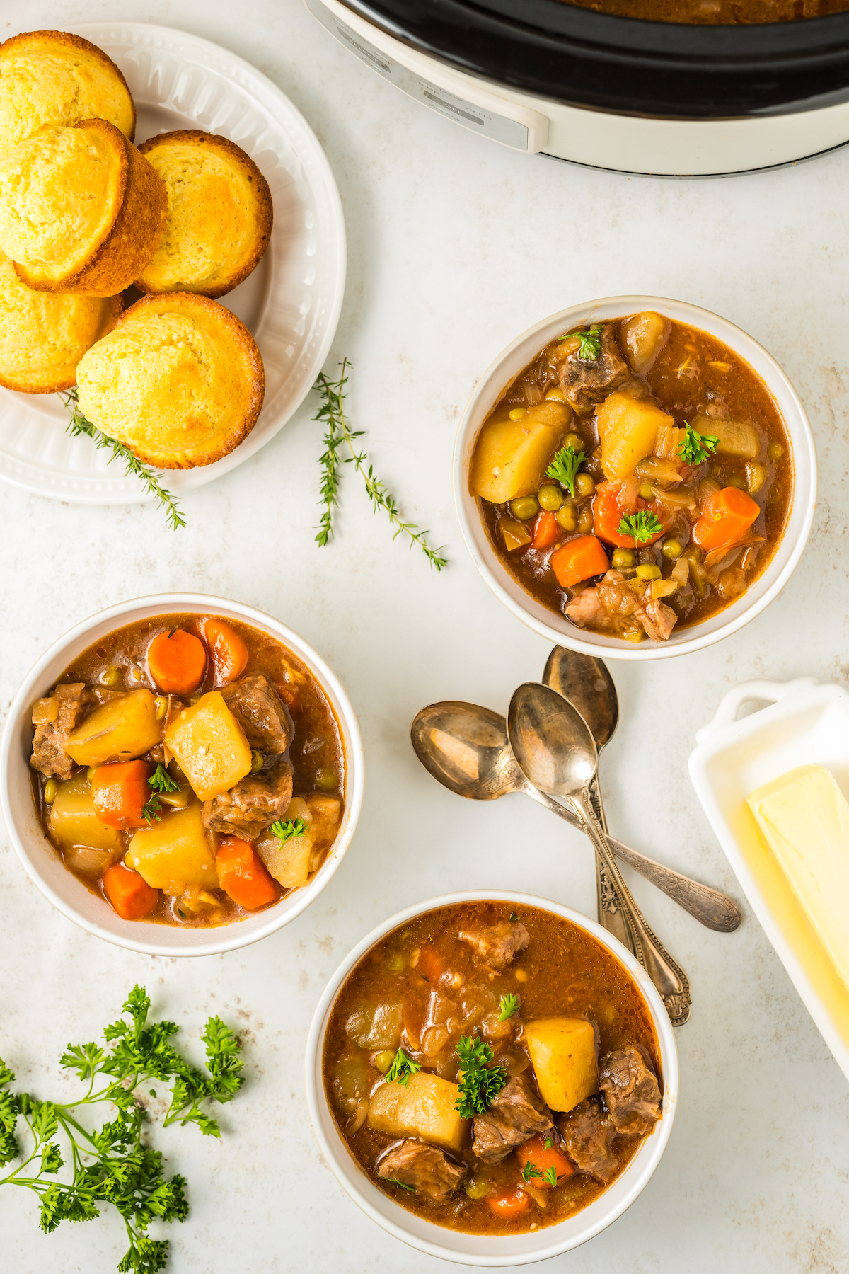 Overhead shot of corn muffins, beef stew, and spoons on a table with fresh herbs.