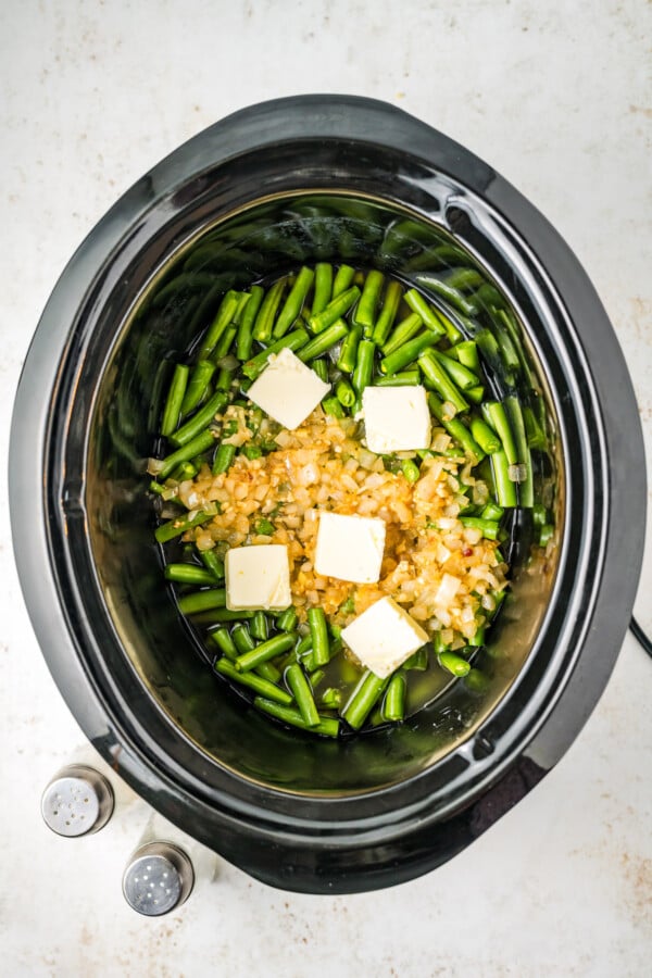 Pats of butter and other ingredients added to green beans in a crock pot.
