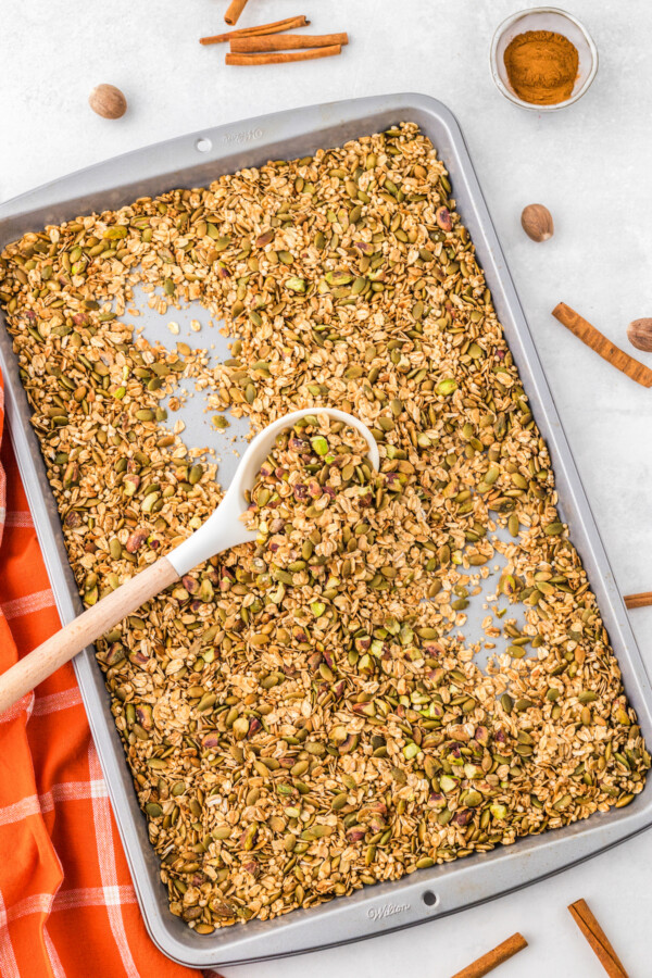 A wooden spoon resting in a sheet pan filled with pumpkin seed granola.