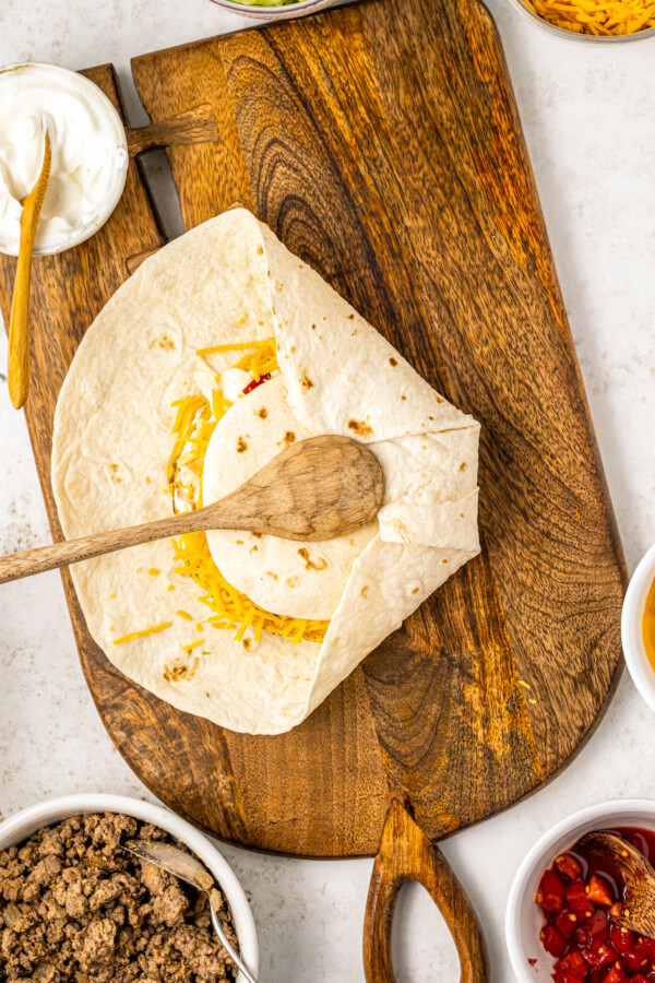 The largest flour tortilla is folded over the smaller tortilla with a wooden spoon being used to hold down the tortillas.