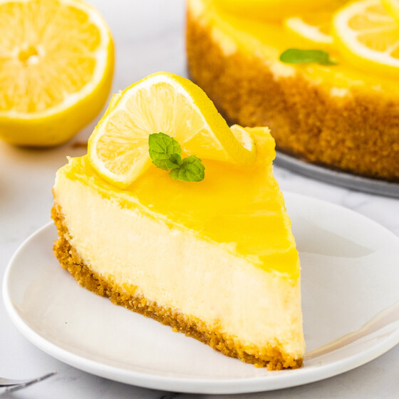 A slice of lemon cheesecake topped with lemon curd and a slice of lemon and mint, sit on a plate.