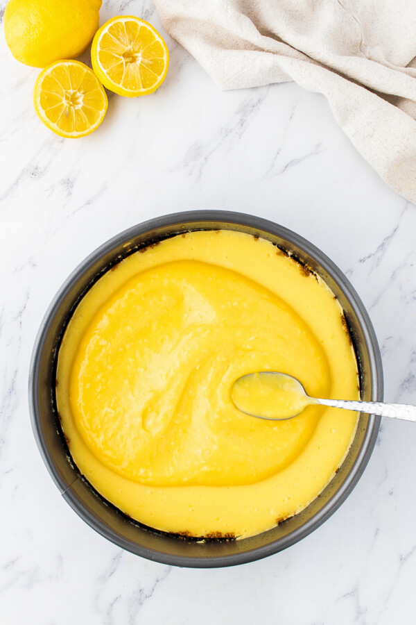 Lemon curd is being spread over cooled cheesecake with a spoon.