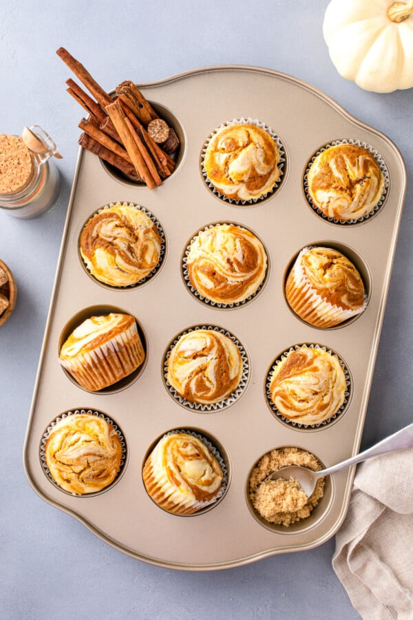 Overhead view of pumpkin cheesecake muffins in a muffin tin with cinnamon sticks and brown sugar on the sides.