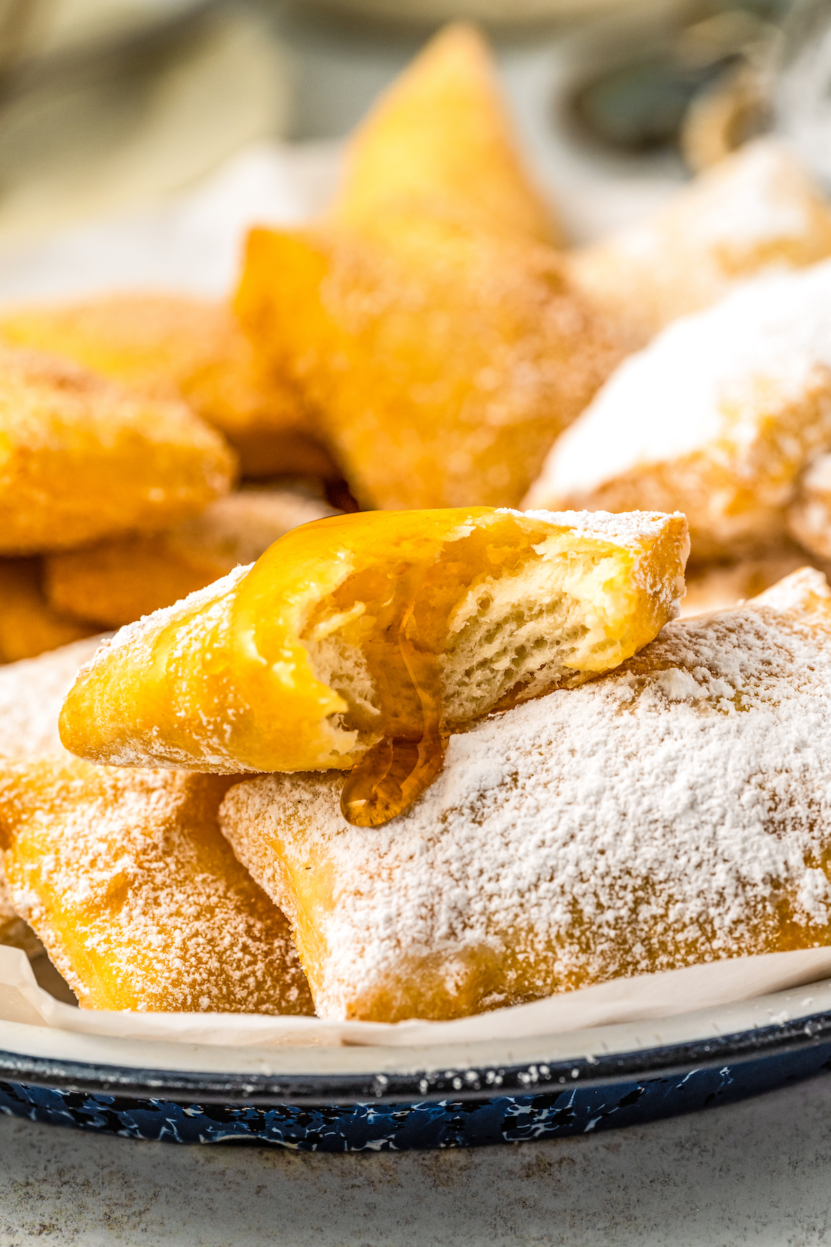 Sopapillas on a plate coated in powdered sugar with honey drizzled over a sopapilla with a bite taken out of it.