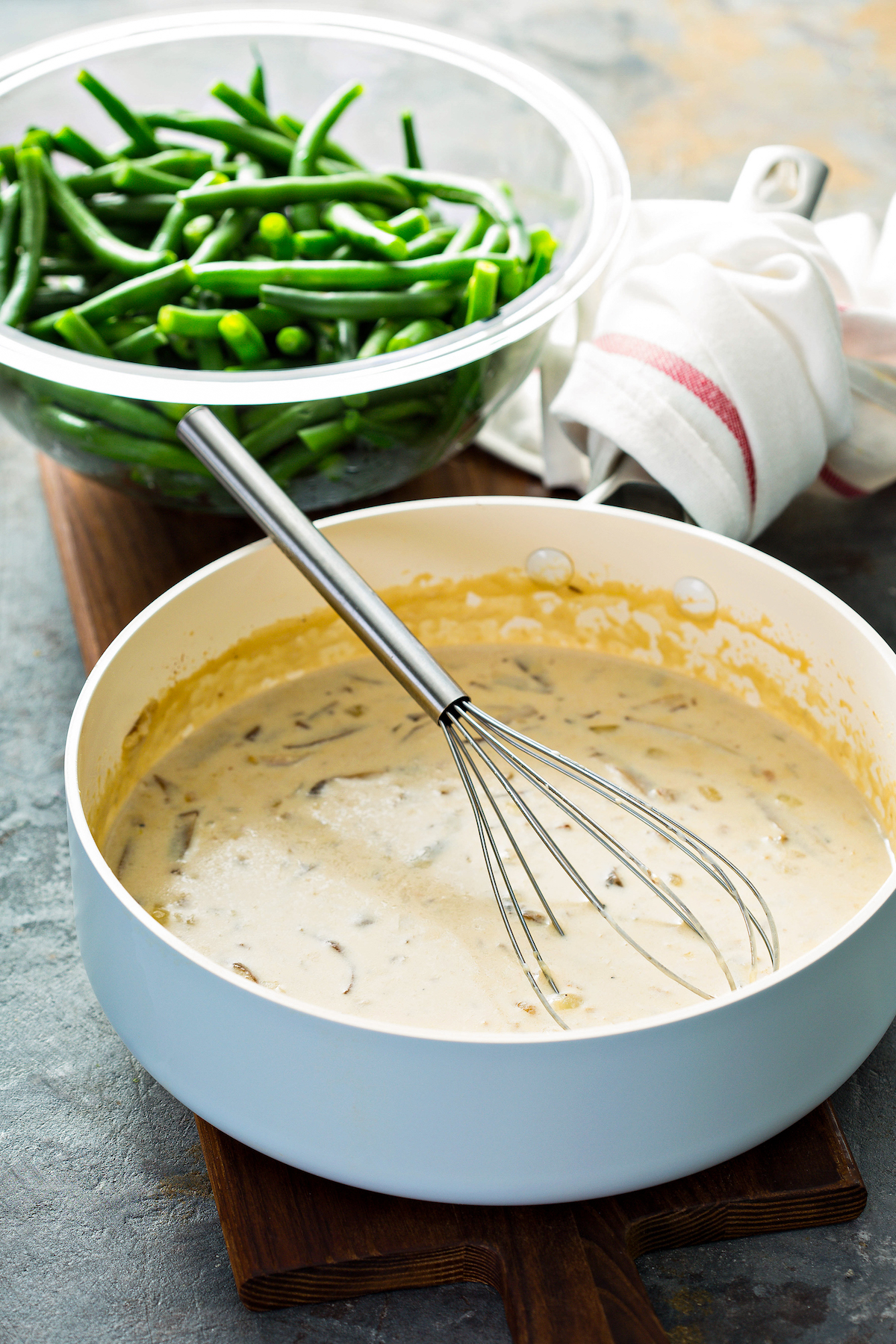 A skillet filled with homemade cream of mushroom soup with a whisk stirring the sauce and a bowl of blanched green beans in the background.