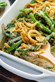 Homemade green bean casserole with fresh green beans and crispy fried onions on top in a baking dish with a wooden spoon scooping out a serving.