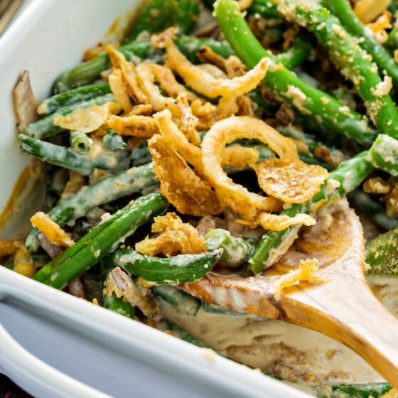 Homemade green bean casserole with fresh green beans and crispy fried onions on top in a baking dish with a wooden spoon scooping out a serving.