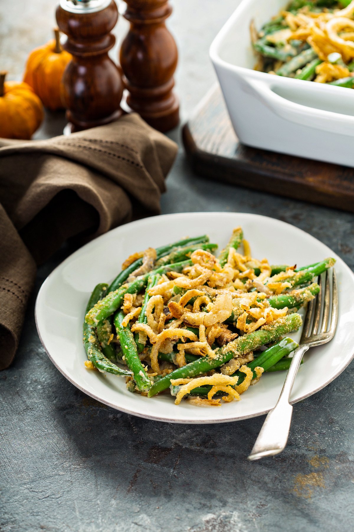 A serving of fresh green bean casserole on a plate with a fork and the whole casserole in a baking dish in the back ground.