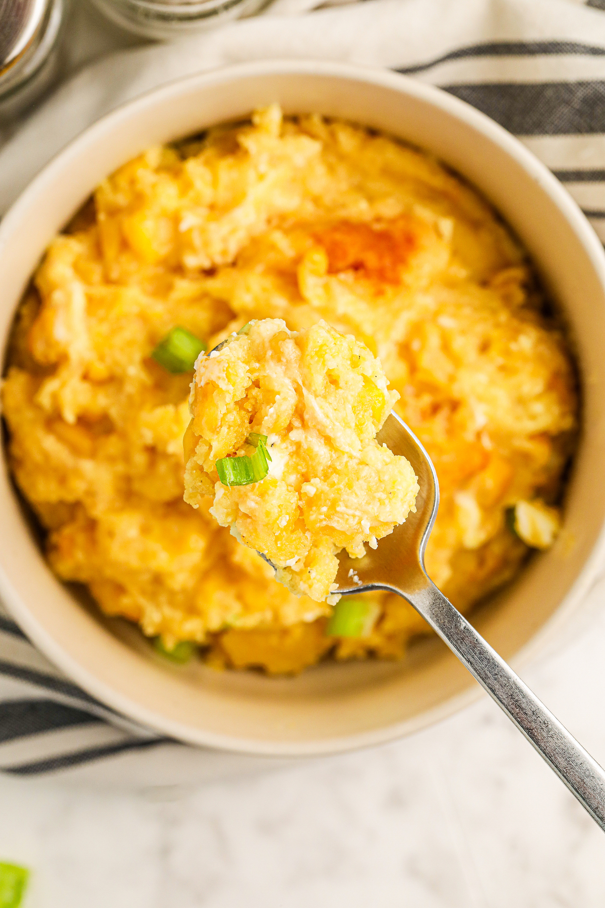 A bowl of sweet corn pudding with chopped green onions on top and a spoon scooping out a bite.