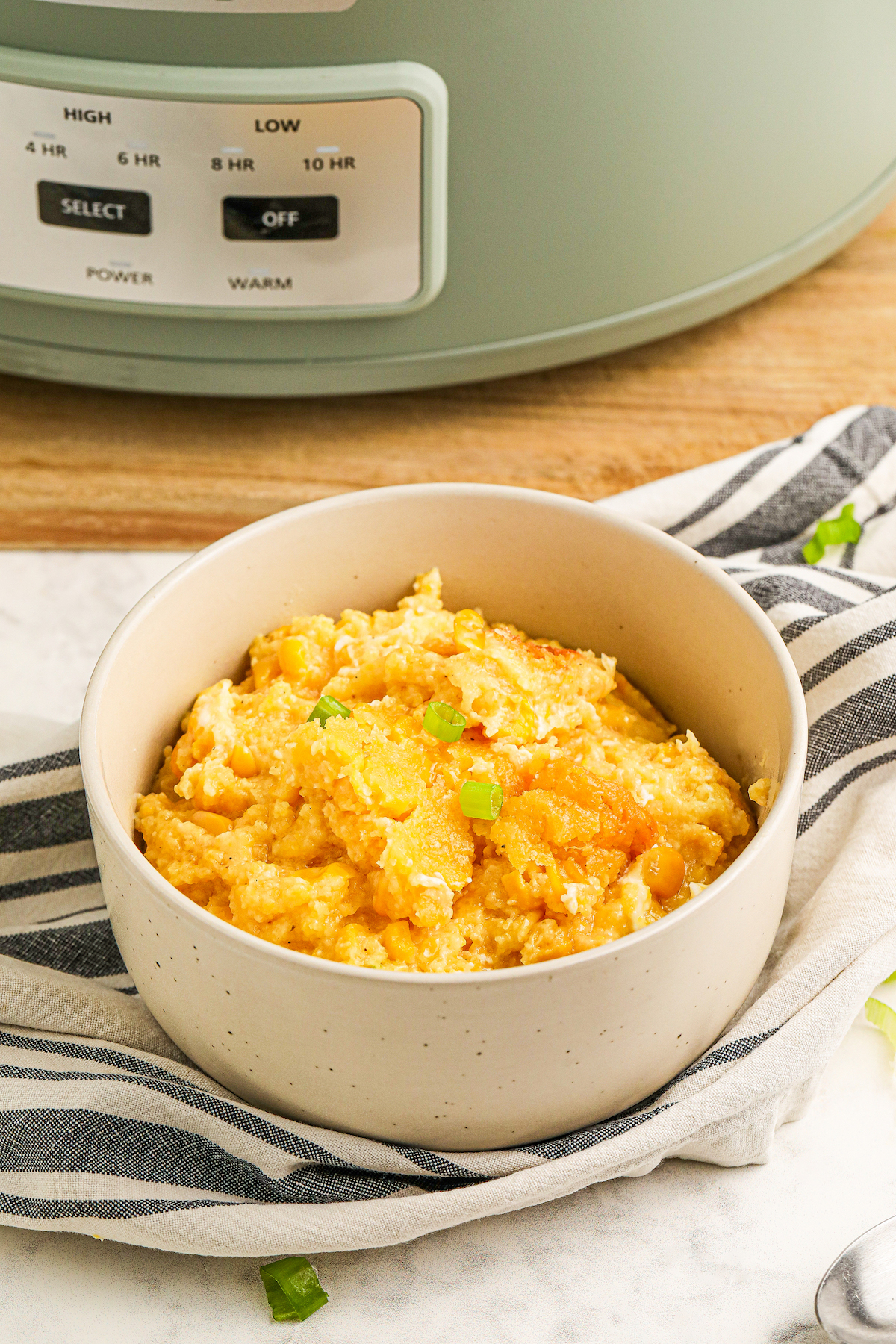 Bowl of sweet and creamy crockpot corn casserole in front of a slow cooker set on a cutting board.
