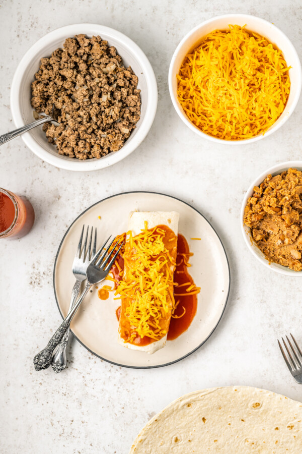 Ingredients for an enchirito in bowls surrounding a plate with a whole enchirito on it with sauce and cheddar cheese on top.
