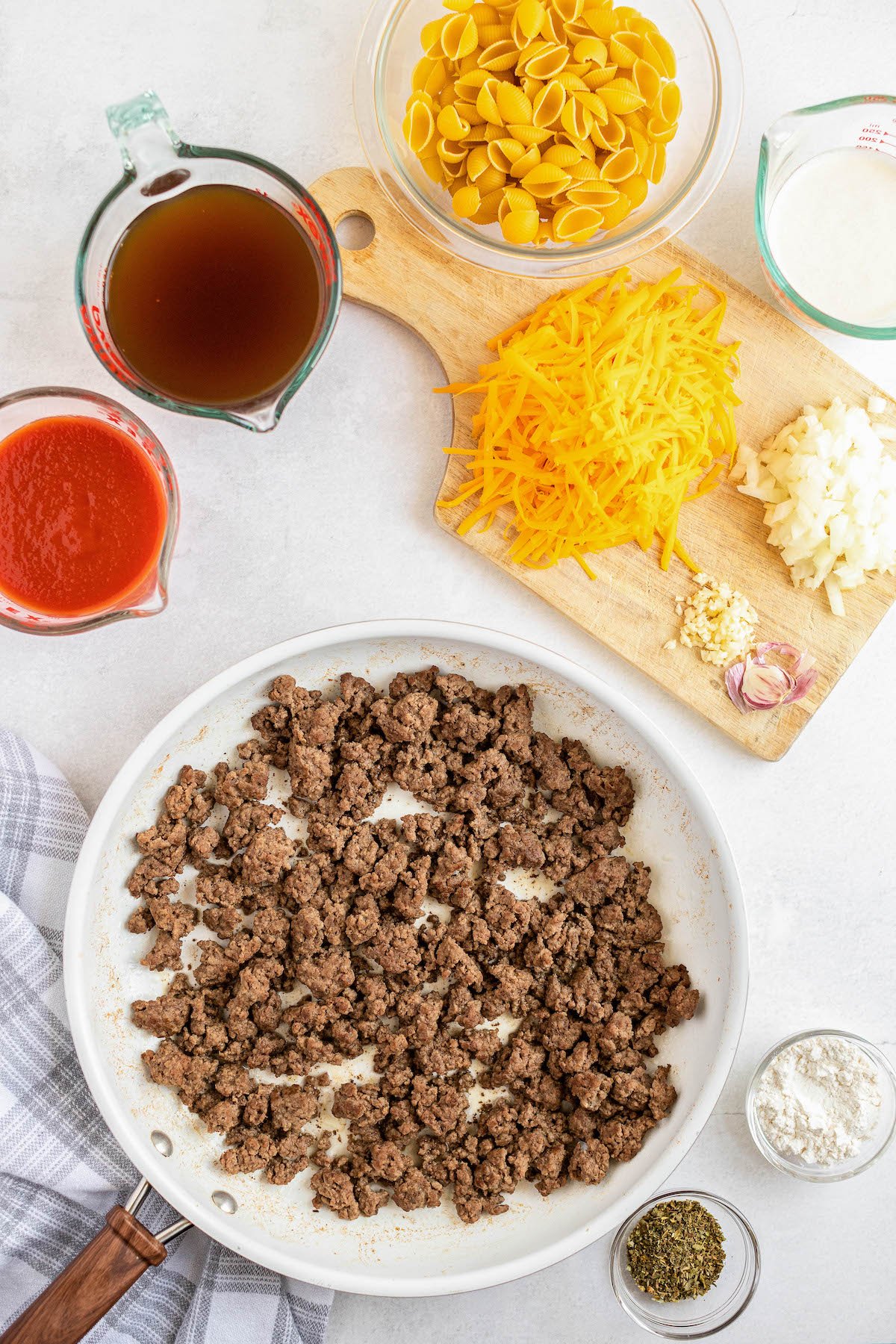Ingredients for homemade hamburger helper recipe arranged on a countertop in bowls with the ground beef in a skillet.