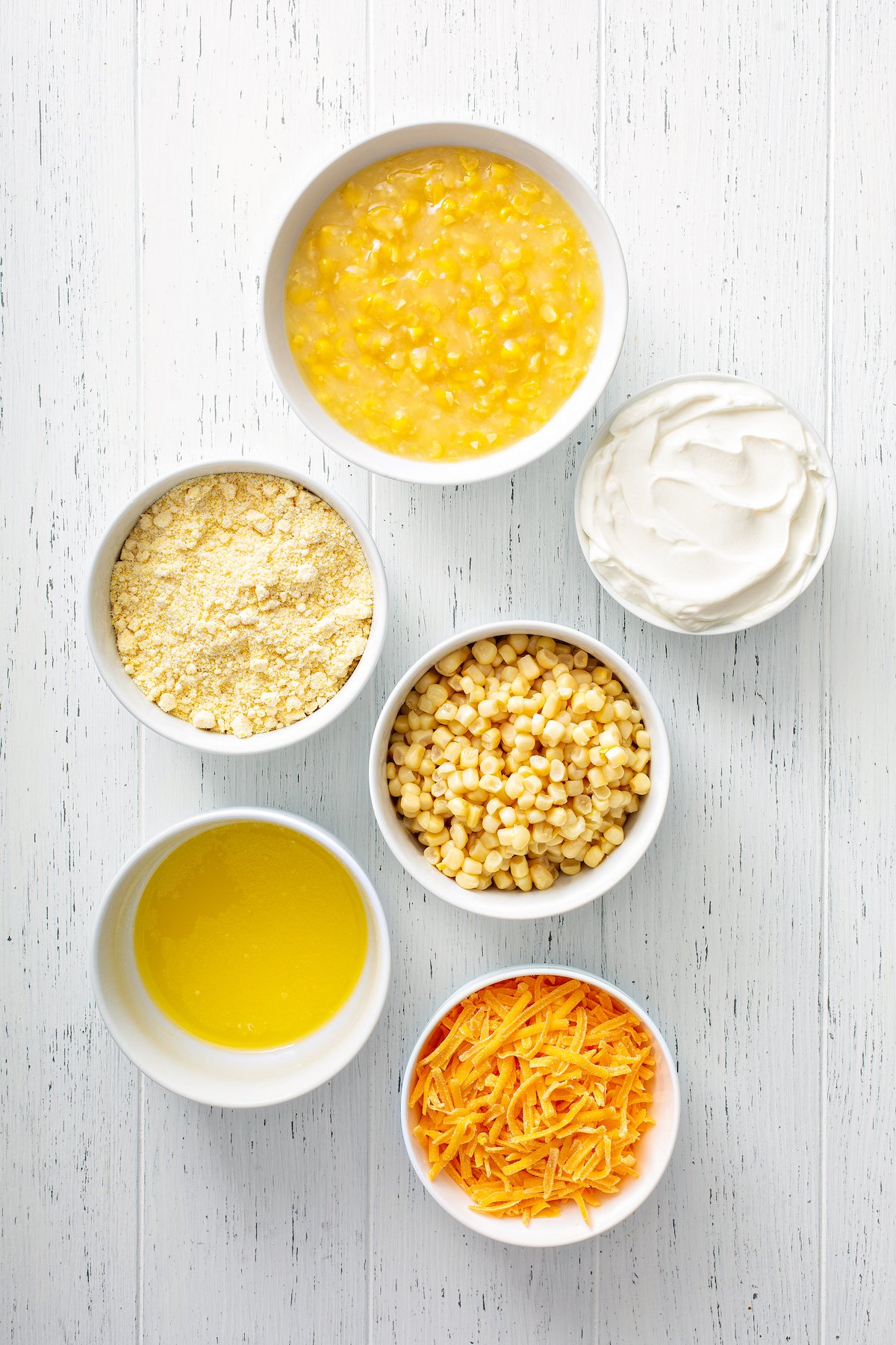 Ingredients for corn casserole recipe arranged in white bowls. From top: a bowl of creamed corn, a bowl of sour cream, a bowl of jiffy cornbread mix, a bowl of frozen corn kernels, a bowl of melted butter and a bowl of shredded cheddar cheese.