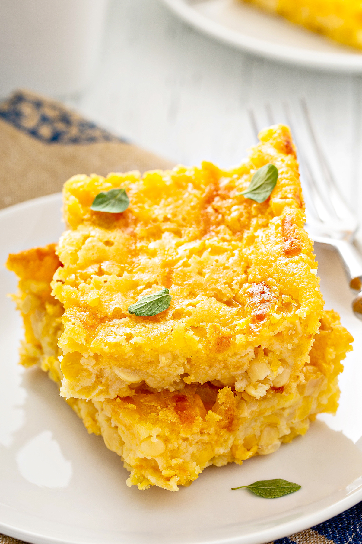 Two squares of soft creamed corn casserole stacked on top of each other on a white plate with fresh herbs sprinkled on top.