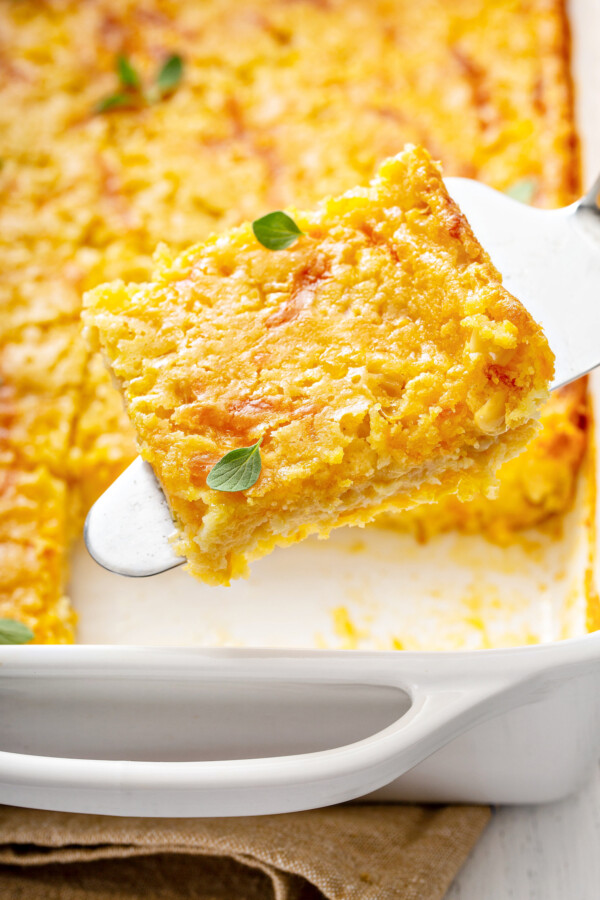 A slice of jiffy corn casserole being lifted out of a white baking dish filled with corn pudding.